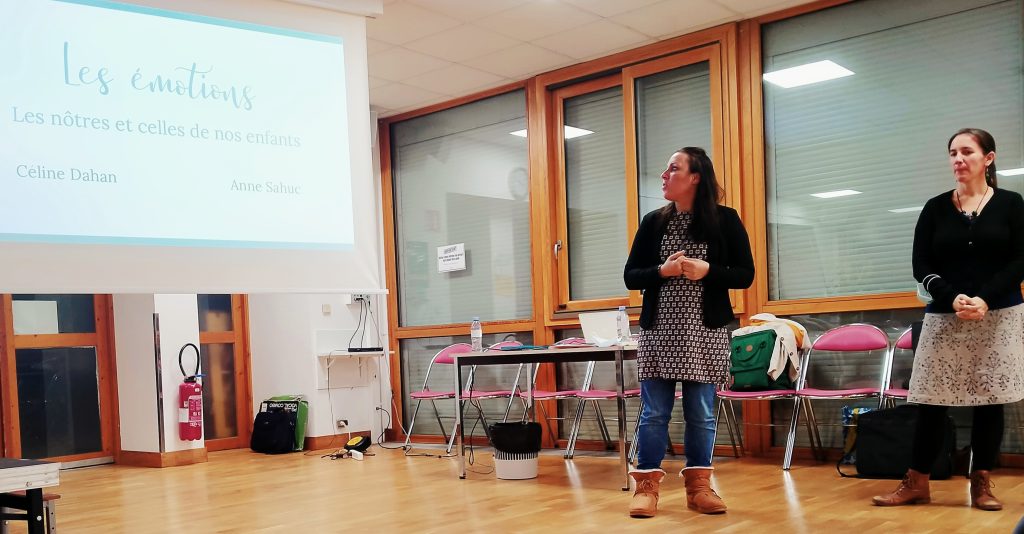 Céline Dahan debout dans une salle. Elle fait une présentation devant un rétroprojecteur. Le titre de l'atelier est: "Les émotions : Les nôtres et celles de nos enfants". À sa gauche une femme se tient elle aussi debout, il s'agit possiblement de Anne Sahuc.