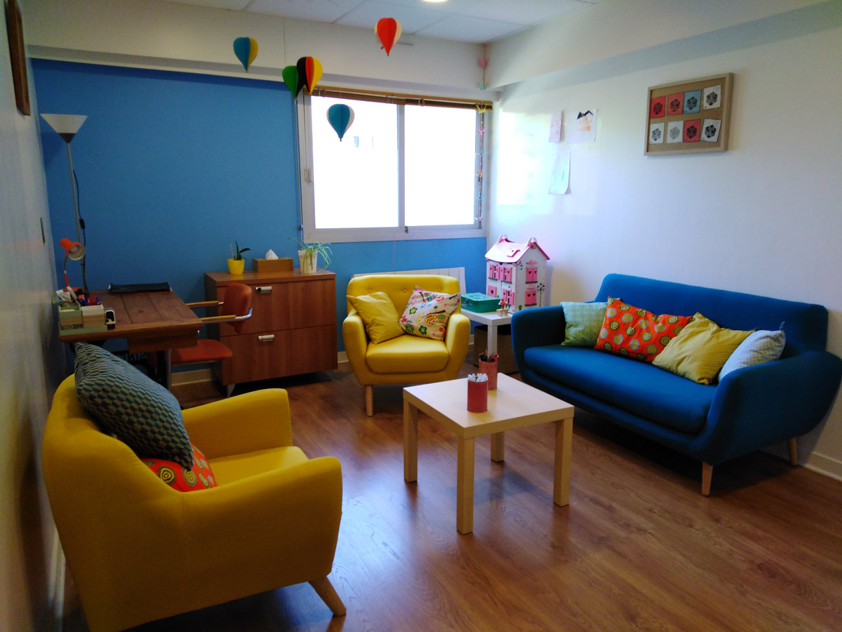 Bureau de psychologue de Céline Dahan. Il y a deux fauteuils jaunes avec des coussins et un canapé bleu, lui aussi avec des coussins, une table basse se tient au milieu.